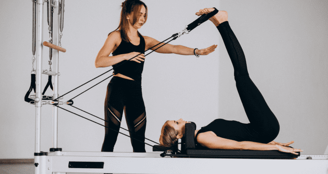 A woman wearing black Pilates wear and doing a reformer and an instructor