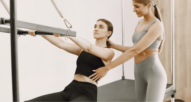 A woman does Pilates in a Cadillac while having her stomach supported by an instructor